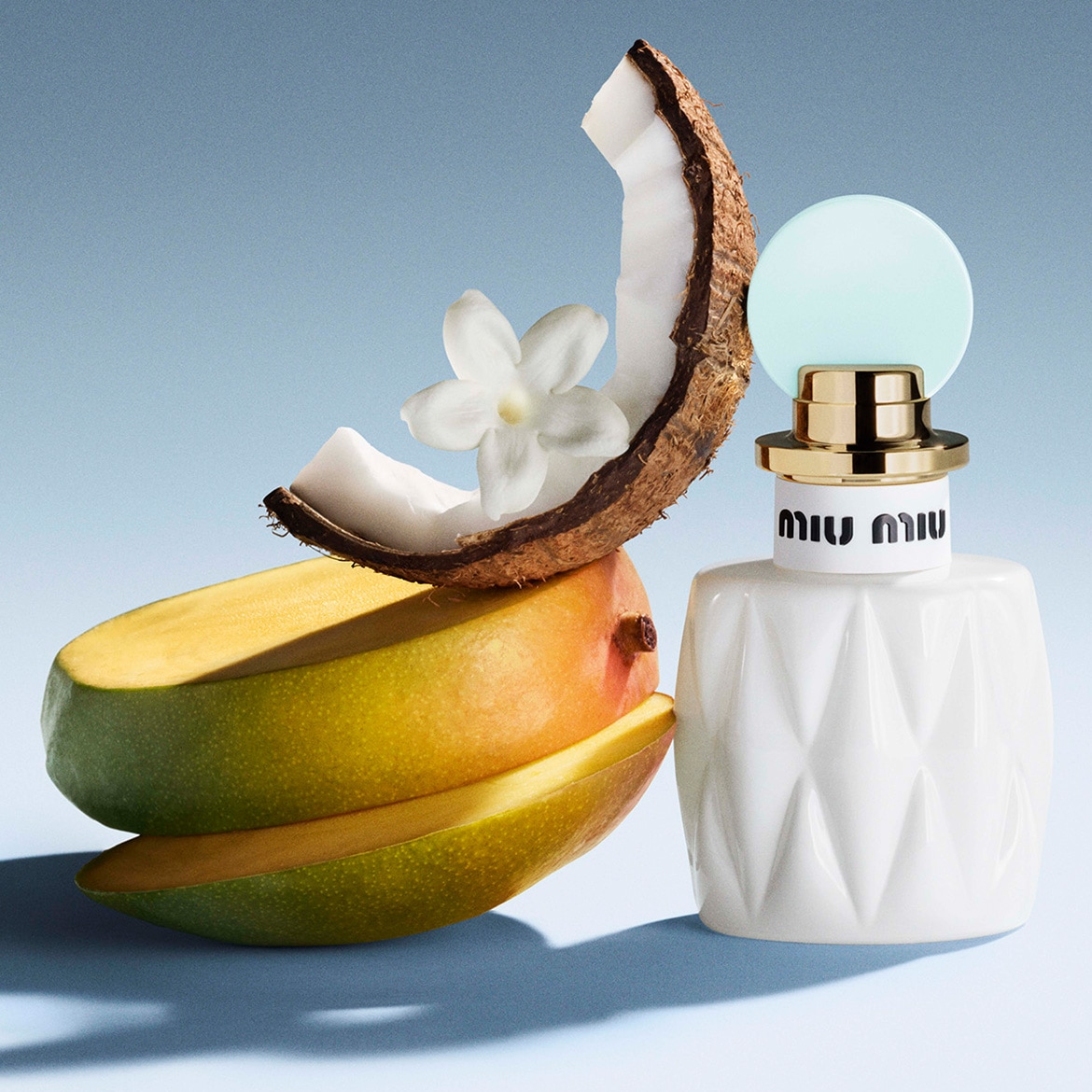 Een witte Miu Miu parfumfles staat naast een stapel mango, kokosnoot en een witte bloem, wat de tropische geur van het parfum benadrukt.