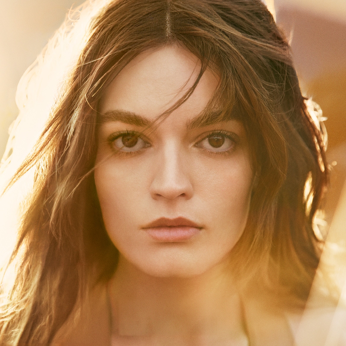 Een close-up van een vrouw met bruin haar en een natuurlijke make-up look, haar gezicht is verlicht door een warme, gouden gloed.